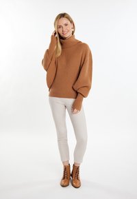 Pulover bej cu guler înalt și nervuri, cu mâneci ample, asortat cu pantaloni bej mulați și cizme scurte din piele maro cu șireturi. Fundal deschis.