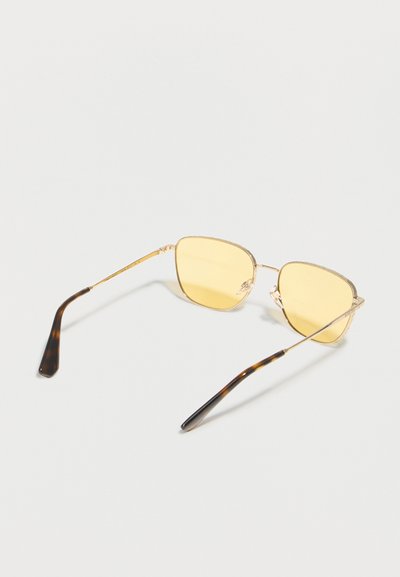 Γυαλιά ηλίου με χρυστό μεταλλικό σκελετό, κίτρινους τετραγωνισμένους φακούς και μύτες ναών με σχέδιο χελώνας πάνω σε λευκή επιφάνεια.