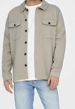 Mand iført let beige skjortejakke med knapper over en hvid t-shirt og lyseblå jeans, med synlig tatovering på venstre hånd.