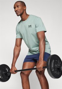 Világoszöld pamut póló fekete Under Armour logóval, tengerészkék rövidnadrággal párosítva. Az egyén egy fekete súlyzót emel.