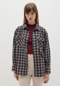 Camisa de franela a cuadros en negro y beige, con mangas largas, dos bolsillos en el pecho y cierre de botones. Se lleva sobre un top corto burdeos, con un ajuste relajado.