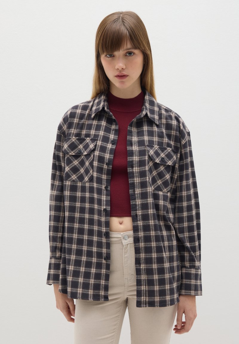 Camisa de franela a cuadros en negro y beige, con mangas largas, dos bolsillos en el pecho y cierre de botones. Se lleva sobre un top corto burdeos, con un ajuste relajado.