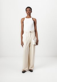 Weißes Tanktop, beigefarbene, hochgeschnittene Hose mit Gürtel und schwarze Ballerinas. Hält eine kleine weiße Handtasche und steht vor einem hellen Hintergrund.