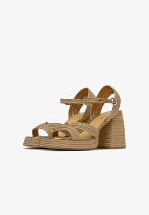 Een paar beige gevlochten sandalen met blokhak, enkelbandjes en zilveren gespen op een witte achtergrond.