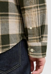 Camisa de franela a cuadros en verde y beige con mangas largas, puños abotonados y dobladillo redondeado, combinada con jeans de mezclilla oscura.