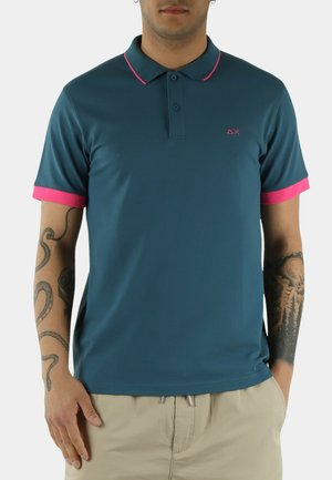 Uomo che indossa una polo verde petrolio scuro con rifiniture rosa sul colletto e sulle maniche, pantaloni beige e tatuaggi visibili su entrambe le braccia.