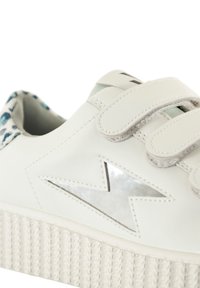 Baskets en cuir blanc avec une semelle en caoutchouc blanc texturée, des sangles Velcro et un accent en étoile argentée réfléchissante sur le côté. Doublure intérieure texturée.