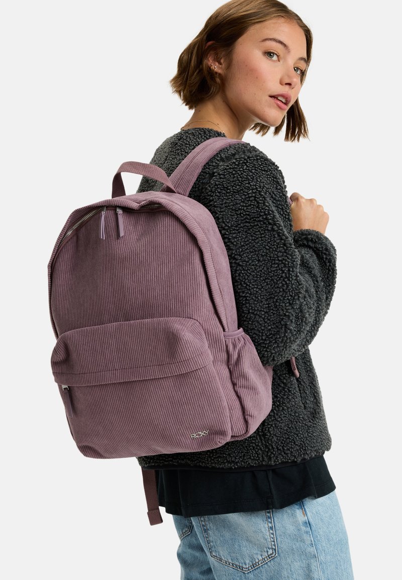 Paarse corduroy rugzak met een textuurafwerking, voorzak en gewatteerde schouderbanden, gedragen over een zwarte fluffy jas en lichtblauwe spijkerbroek.