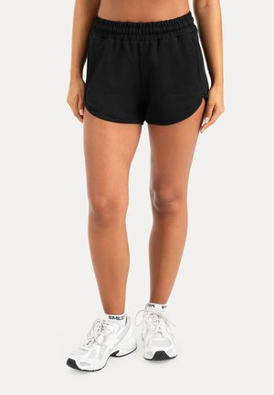 Kvinde iført sorte atletikshorts, hvide mærkede sokker og hvide løbesko står på en ensfarvet baggrund.