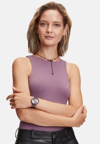 Paarse mouwloze top, gladde textuur. Zwarte horloge met een ronde wijzerplaat, zilveren accenten en een gestructureerde band, gecombineerd met zwarte aansluitende broeken.