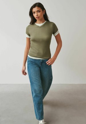 Jeune femme portant un t-shirt vert olive à col en V superposé sur une chemise blanche, associé à un jean bleu et des baskets blanches, debout à l'intérieur.