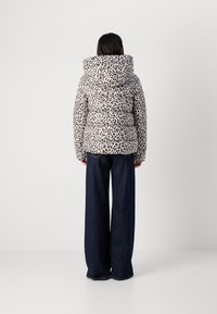 Eine Person steht mit dem Rücken zum Betrachter, trägt eine Steppjacke mit Leopardenmuster und weite dunkle Jeans vor einem einfarbigen Hintergrund.