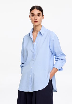 Kobieta o ciemnych włosach w luźnej, jasnoniebieskiej koszuli w kratę, z podwiniętymi rękawami, stojąca z jedną ręką w kieszeni na białym tle.