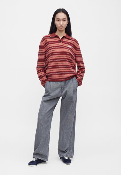 Femme aux longs cheveux foncés portant un polo à manches longues rayé rouge et rose, un pantalon large gris et des baskets bleu marine, debout les mains dans les poches.