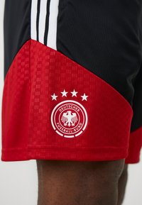 Κόκκινα και μαύρα αθλητικά σορτς με υφή, διακοσμημένα με τρεις λευκές ρίγες και το λογότυπο της Deutscher Fußball-Bund με τέσσερις αστέρες.