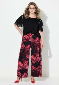 Černý top s průhlednými rukávy a perlovými detaily, spárovaný s širokými květinovými kalhotami v červené a černé barvě. Doplněno červenými vysokými podpatky a oversized slunečními brýlemi.