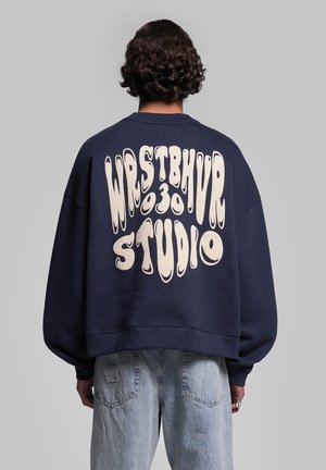 Persona con capelli corti e ricci indossa una felpa blu navy con "WRSTBHVR 930 STUDIO" in grandi lettere bianche distorte sulla schiena.