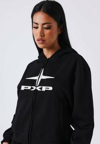 Haut à capuche zippé noir en tissu doux, avec un logo graphique blanc sur le devant, une capuche avec cordon de serrage et des poches latérales.