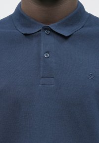 Polo navy bleu en tissu texturé, avec une patte de boutonnage à deux boutons, un col et un petit logo brodé sur la poitrine.