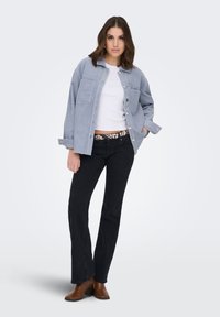 Naine seisab ühe käega taskus, kannab helehalli kopprijope, valget lühikest toppi, musta laiade säärtega teksapükse ja pruune pahkluu saapaid.