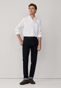 Camisa blanca de botones, jeans oscuros de mezclilla y zapatos marrones. La camisa tiene un bolsillo en el pecho; los jeans tienen costuras claras y puños enrollados.