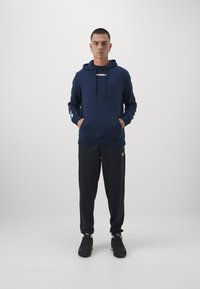 Sweat à capuche bleu marine avec cordons, poche avant et logo blanc ; associé à un pantalon de sport noir et des baskets noires. Design simple.