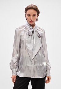 Shiny silver blouse with bow tie detail at neckline, long sleeves, gathered shoulders, and a slightly loose fit. Smooth, reflective texture.