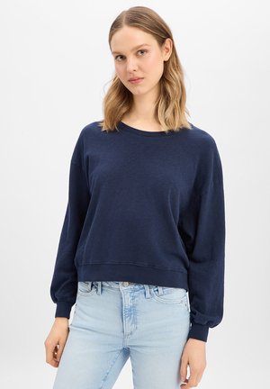 Junge Frau mit schulterlangem blondem Haar, die ein marineblaues Langarm-Sweatshirt und hellblaue Jeans trägt, vor einem schlichten Hintergrund stehend.