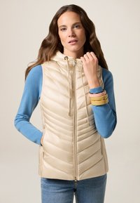 Beige gesteppte Weste mit Kapuze, silbernem Reißverschluss und Zickzackmuster. Getragen über einem blauen Langarmoberteil, kombiniert mit übereinander getragenen Armbändern.