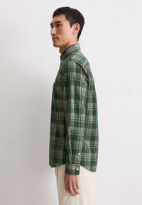 Groene geruite overhem met lange mouwen, knoopsluiting aan de voorkant en een zak. Gemaakt van een gestructureerde stof met een lichter groen en crèmekleurig ruitpatroon.