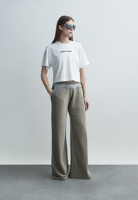 Donna che indossa pantaloni grigi oversize, t-shirt bianca corta e grandi occhiali da sole riflettenti, in piedi davanti a uno sfondo semplice di colore grigio chiaro.