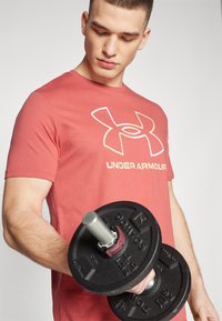 Rød t-skjorte med korte ermer og hvit Under Armour-logo, holder en svart vektplate med sølvhåndtak, som viser en muskuløs arm.