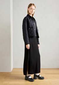 Black quilted cropped jacket with a collar, worn over a black dress featuring a side slit. Black heeled shoes complete the outfit.