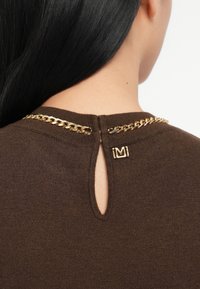 Bruine gebreide top met een keyhole-detail aan de achterkant, accented met een gouden ketting en een klein logo-label bij de halslijn.