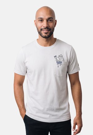 Lächelnder glatzköpfiger Mann mit Bart, der ein weißes T-Shirt mit einer Katze trägt, die einen Pinsel hält und aus der Brusttasche hervorlugt, die Hände in den Taschen.