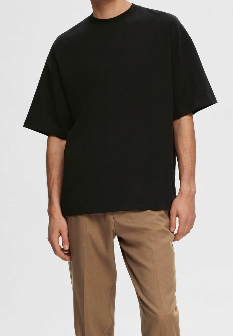 Hombre con una camiseta negra holgada de manga corta y pantalones beige, de pie contra un fondo claro liso.