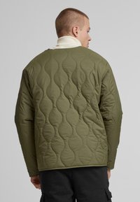 Olivgrüne Steppjacke mit einem Muster aus gewellten Nähten, ausgestattet mit langen Ärmeln und einem Kragen, getragen über einem cremefarbenen Rollkragenpullover.