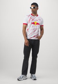 Balta sportinė marškinė su raudonais akcentais, turinti įstrižas linijas ir "Red Bull" logotipą, derinama su juodomis džinsinėmis kelnėmis ir sportiniais bateliais.