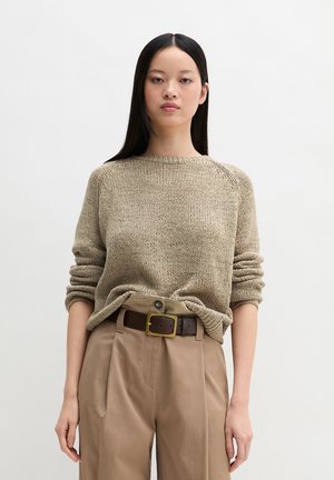 Młoda kobieta z długimi czarnymi włosami, ubrana w beżowy dziergany sweter włożony w spodnie z wysokim stanem w kolorze tan, z ciemnobrązowym paskiem.