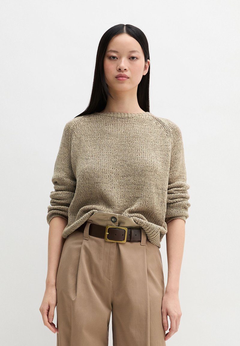 Giovane donna con lunghi capelli neri che indossa un maglione beige a maglia infilato in pantaloni color cammello a vita alta con una cintura marrone scuro.