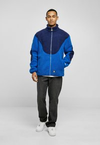 Blue and navy fleece jacket with high collar, full zipper, and two side pockets; textured fabric with a contrasting color panel design.