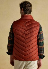 Bordová nafúknutá vesta s chevronovým šitím, ľahká tkanina, vysoký golier a hladká textúra, nosená cez károvanú flanelovú košeľu.