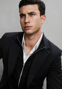 Outfit formale con un blazer nero, un maglione zip nero sopra una camicia bianca. Tessuto liscio, vestibilità sartoriale, con un design moderno e minimalista.