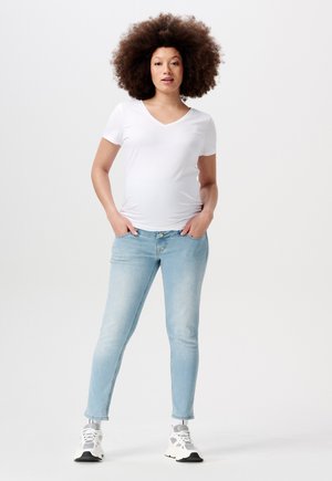 Witte V-hals t-shirt gecombineerd met lichtblauwe skinny jeans. Model staat met handen in de zakken, draagt witte en grijze sneakers. Effen achtergrond.