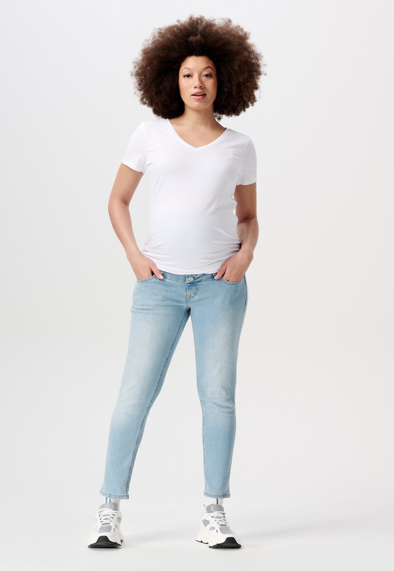 Witte V-hals t-shirt gecombineerd met lichtblauwe skinny jeans. Model staat met handen in de zakken, draagt witte en grijze sneakers. Effen achtergrond.