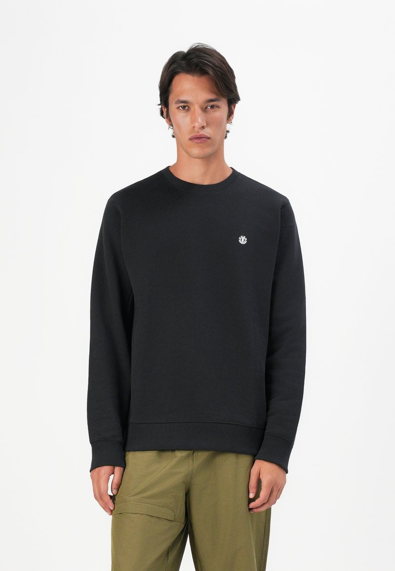 Giovane uomo che indossa un pullover nero a girocollo con un piccolo logo sul petto e pantaloni cargo verde oliva, in piedi davanti a uno sfondo bianco.