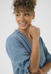 Jasnoniebieska lniana bluzka z falbaniastym dekoltem i mankietami, charakteryzująca się luźnym krojem i subtelną fakturą. Modelka uśmiecha się, pokazując ramię.