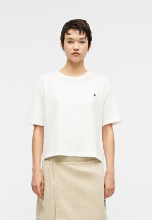 Rövid sötét hajjal rendelkező fiatal áll egy világos, egyszínű háttér előtt, laza, fehér rövid ujjú felsőt és bézs szoknyát viselve.