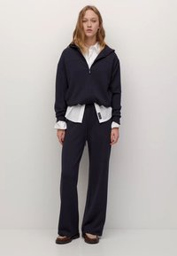 Marineblå hættetrøje med lynlås og høj krave, kombineret med en hvid skjorte med knapper og løstsiddende marineblå bukser. Glat tekstur, afslappet design.