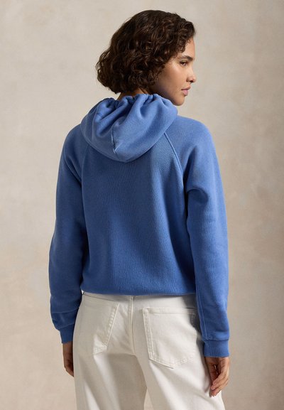 Jaunas asmuo su garbanotais plaukais, dėvintis mėlynę su gaubtu ir baltas kelnes, stovintis nugara į priekį, galva pasukta į šoną.